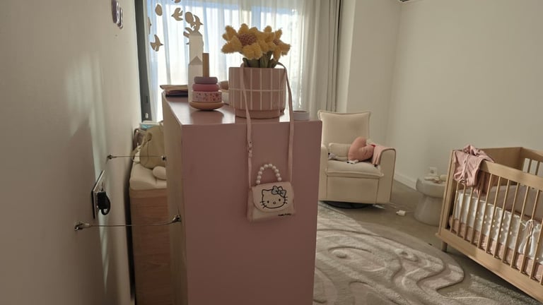A modern nursery room with a large mauve cube structure decorated with dried flowers, a Hello Kitty purse, and a crib visible in the background.